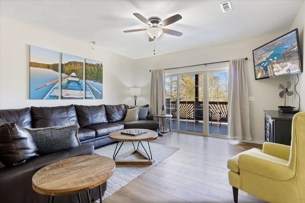A cozy living room with a leather sectional sofa, wooden coffee tables, large wall art of a lake scene, and sliding glass doors that open to a deck. The layout encourages relaxation, socializing, and enjoying scenic views, with easy access to the outdoors.