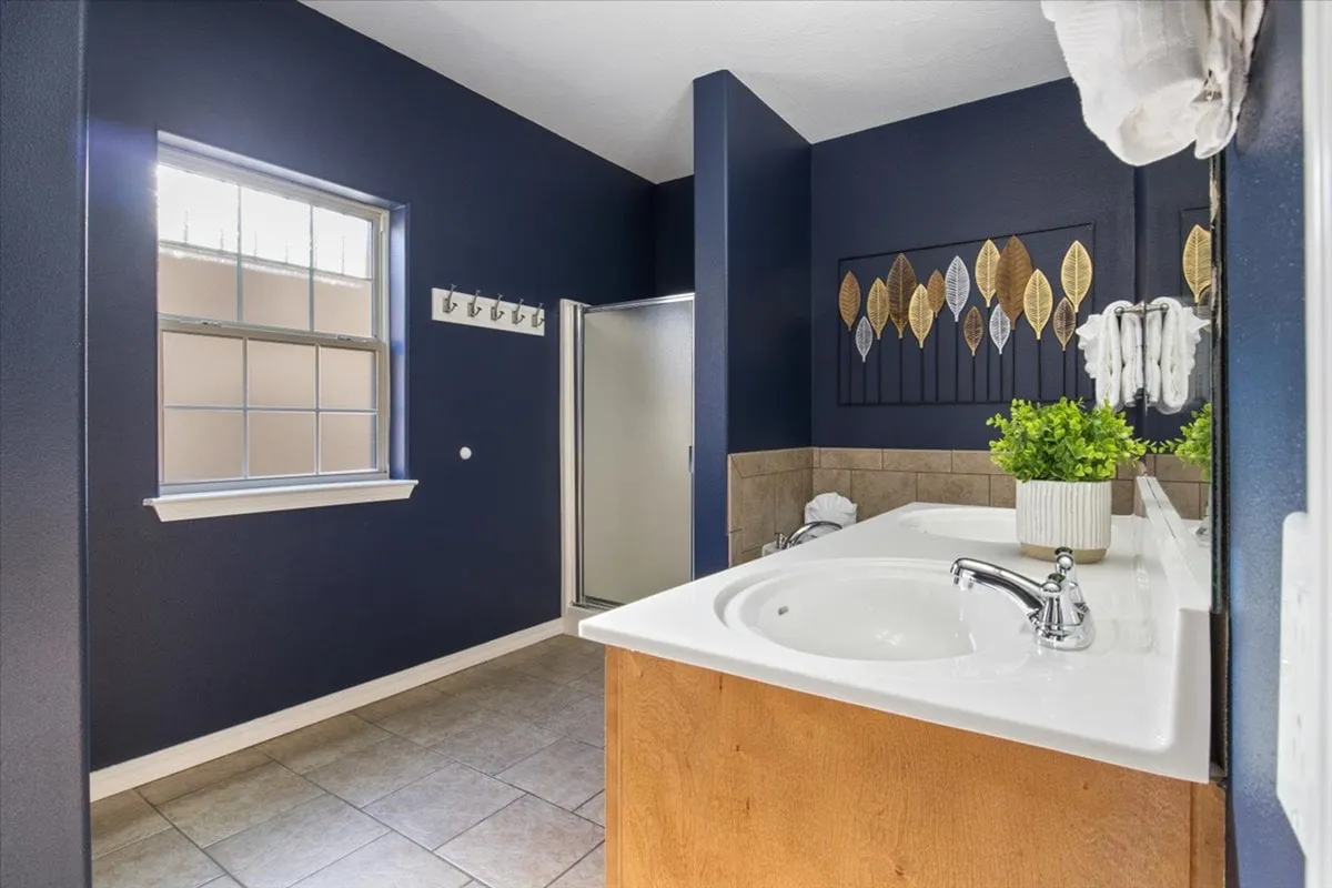 A modern bathroom with dual sinks, leaf-themed artwork, a green plant for a touch of nature, and a tranquil ambiance. The space suggests comfort and space for personal care routines.