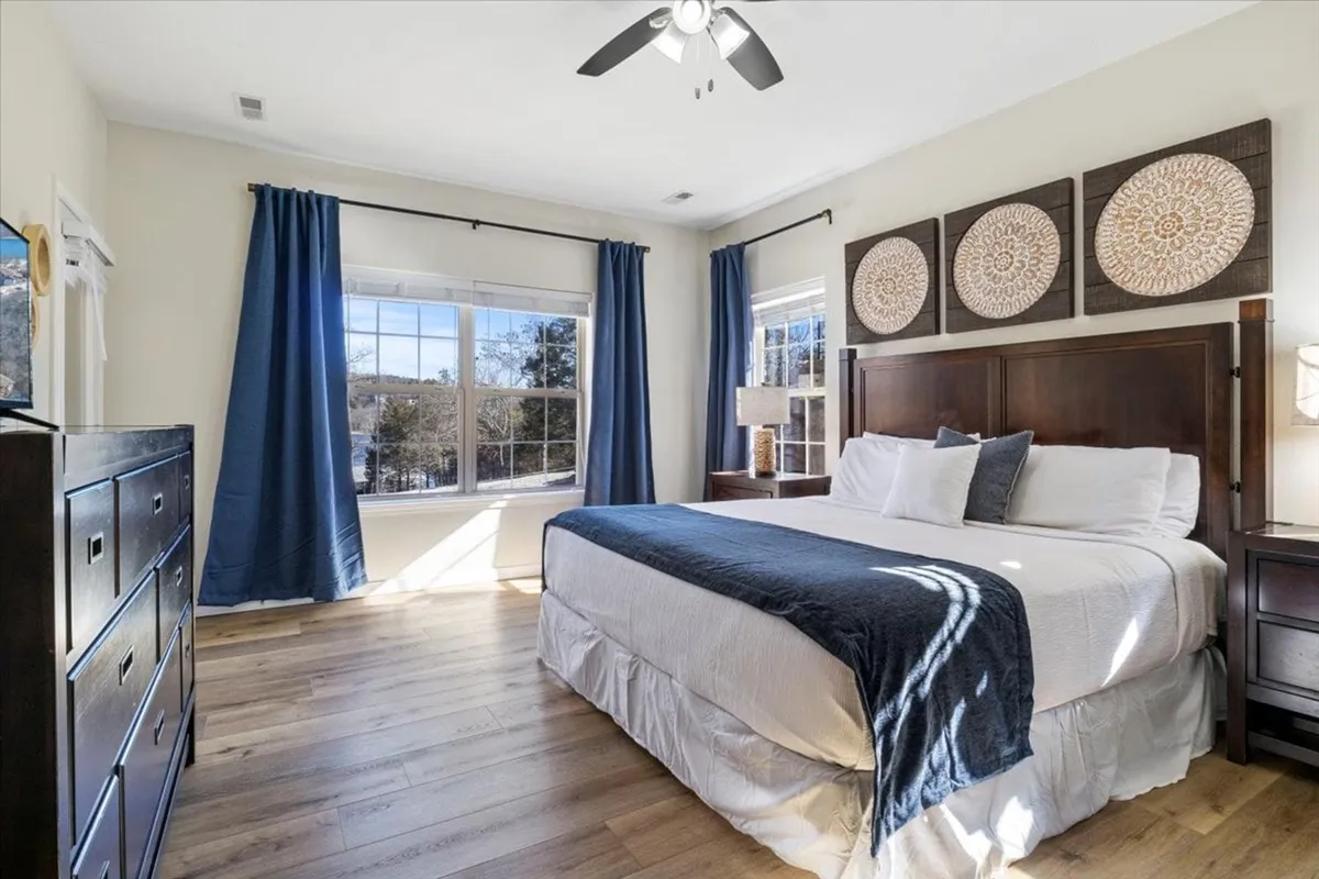 A tranquil bedroom featuring a plush bed with a blue throw, abundant natural light streaming in through a large window, decorative wall art, and a minimalistic aesthetic. The environment feels peaceful and inviting, ideal for rest and relaxation.