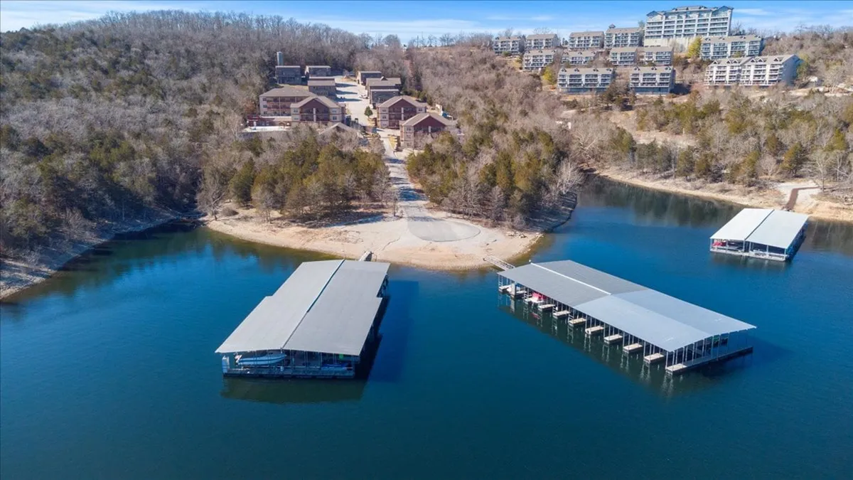 A tranquil lakeside scene featuring docks extending over the water, residential buildings in the background, and a lush tree line. There are visible pathways and open spaces ideal for leisurely walks and outdoor activities. The relaxed atmosphere suggests opportunities for nature exploration and waterfront recreation.