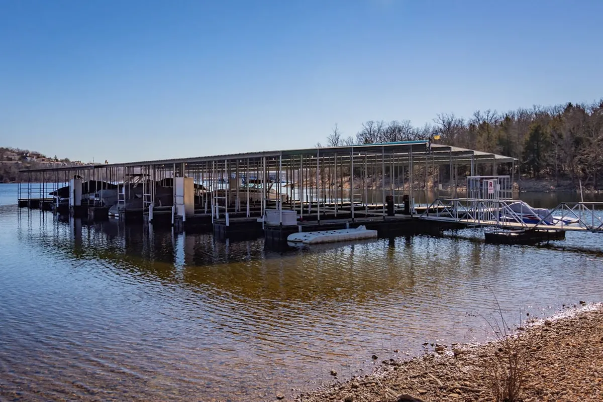 A peaceful waterfront shot with a boat dock extending into the calm lake. Several boats are moored along the dock, emphasizing the ease of launching watercraft. The shoreline is green and inviting, with open skies hinting at beautiful sunsets.