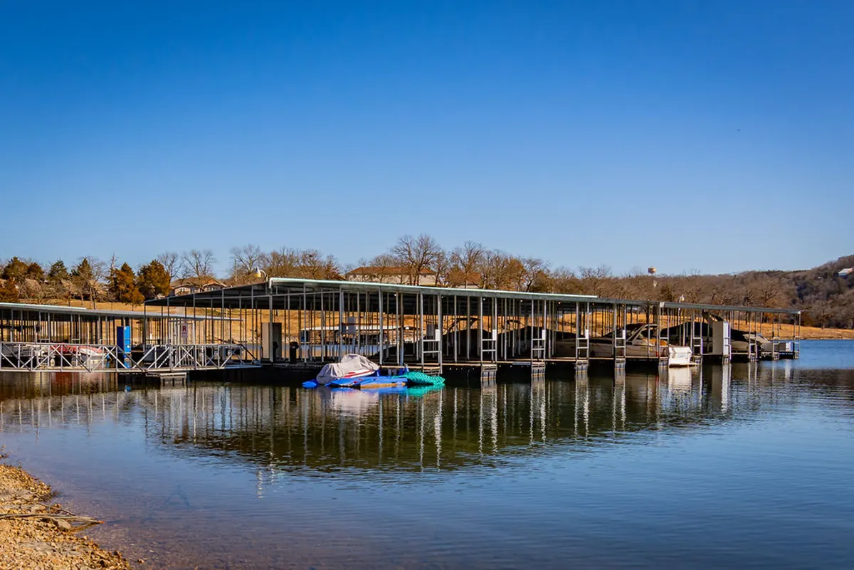 A peaceful lakeside dock scene with boats tied up alongside, surrounded by trees and calm water. The open sky and gentle waves evoke a relaxed, idyllic lakeside lifestyle.