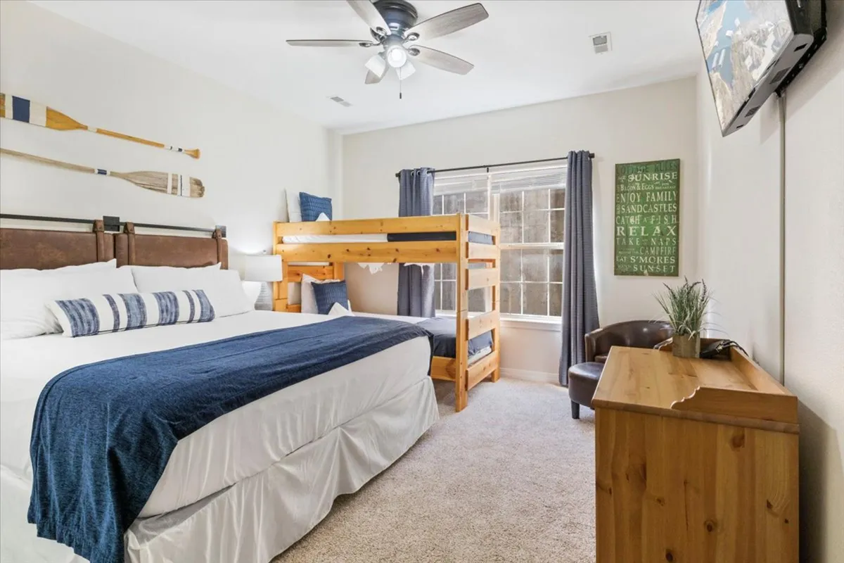 A spacious bedroom with a large king-sized bed dressed in blue and white bedding, a twin bunk bed perfect for children, wooden oars as wall decor, a flat-screen TV mounted on the wall, and a wooden dresser. The layout is designed to comfortably accommodate family groups or multiple guests.