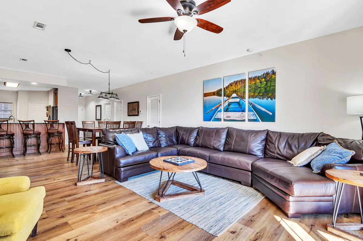 This image highlights a spacious open-concept living area with a large sectional sofa, a dining area with bar stools, and decorative wall art depicting a lake scene. The coffee table and arrangement emphasize a comfortable space for relaxing or entertaining, with ample seating for groups.
